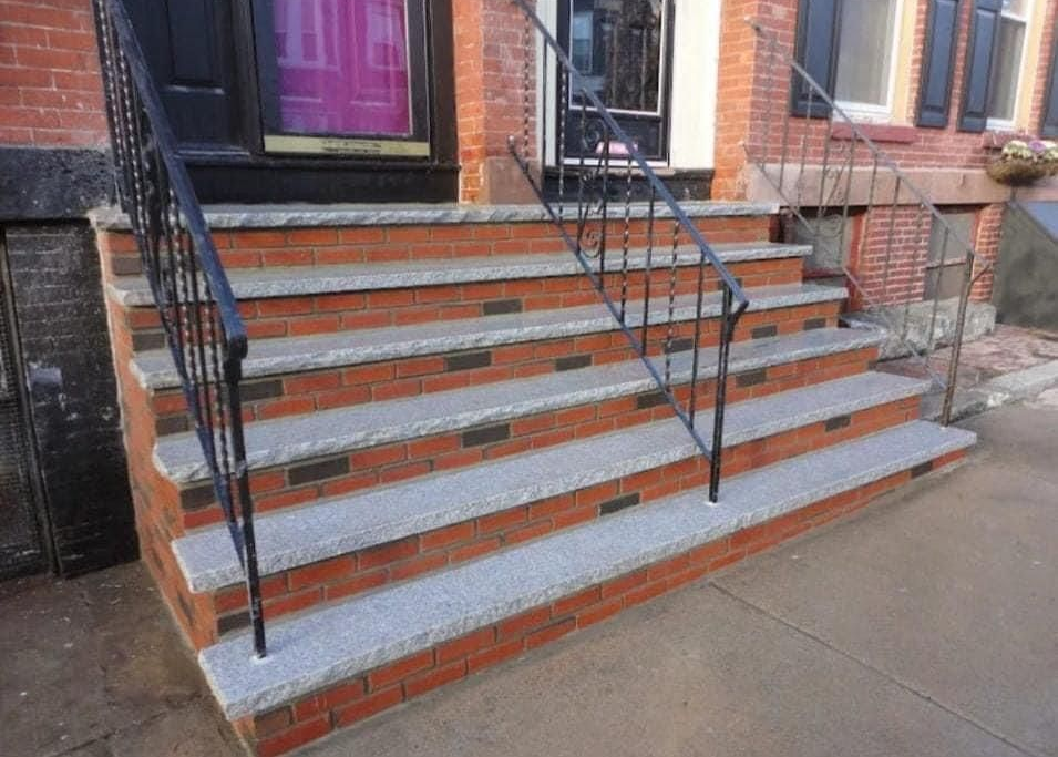 Front steps of a brick building with black railings and gray concrete treads leading to a doorway