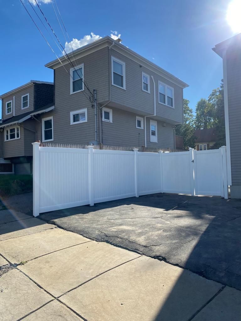 White vinyl fence beside a tan two-story house under a bright blue sky, viewed from a paved driveway.