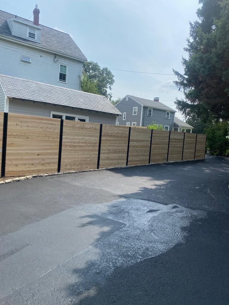 Wooden privacy fence along a driveway beside houses and a large tree.