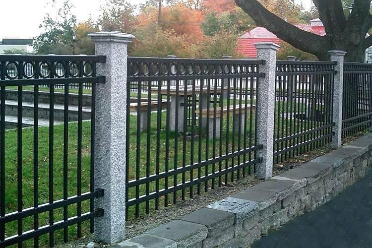 Black metal fence with stone pillars along a sidewalk by a road, with autumn trees in the background