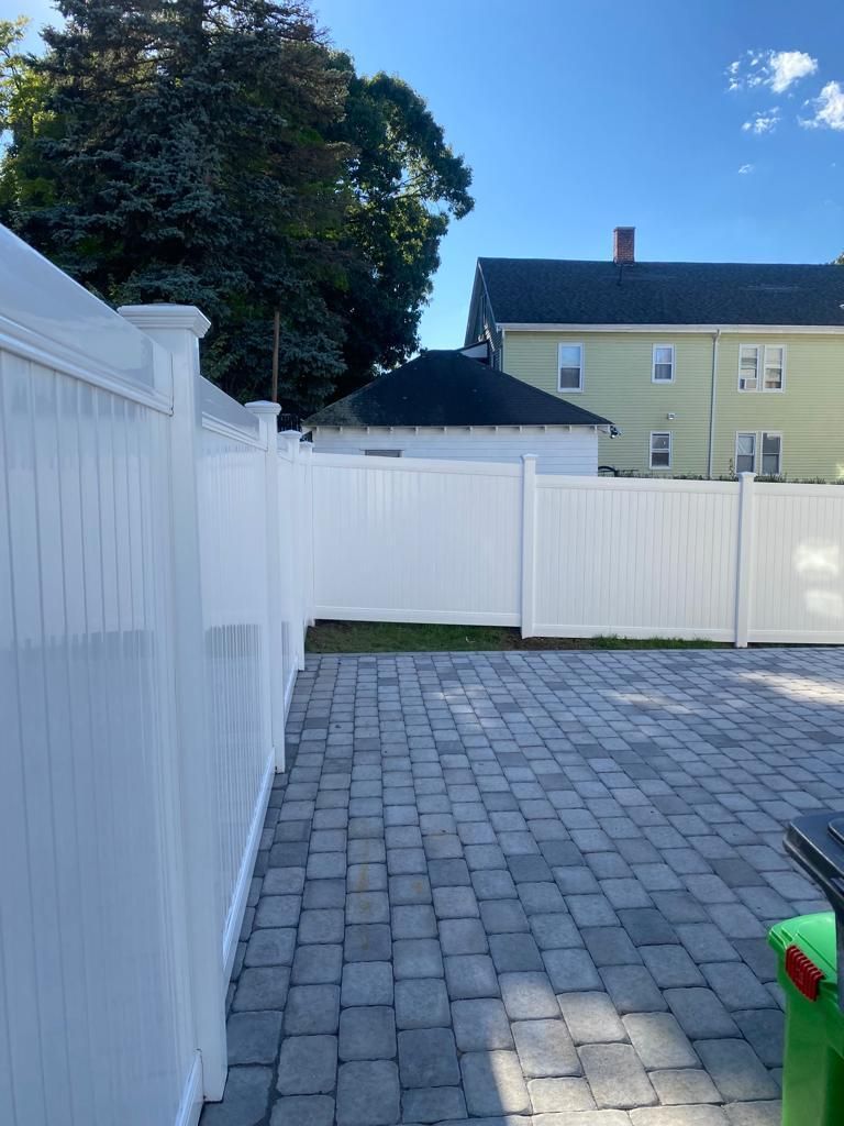White fenced backyard with gray paver patio, tree, and yellow house in background