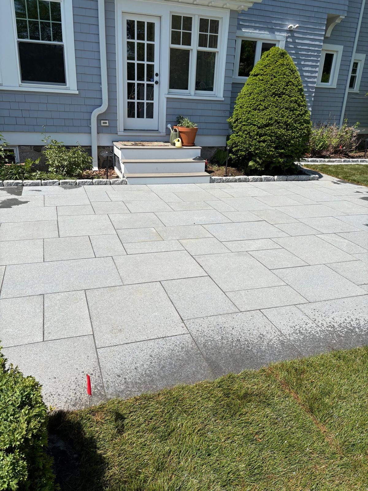 Stone patio leading to a house entrance with white steps, blue siding, and a trimmed evergreen shrub.