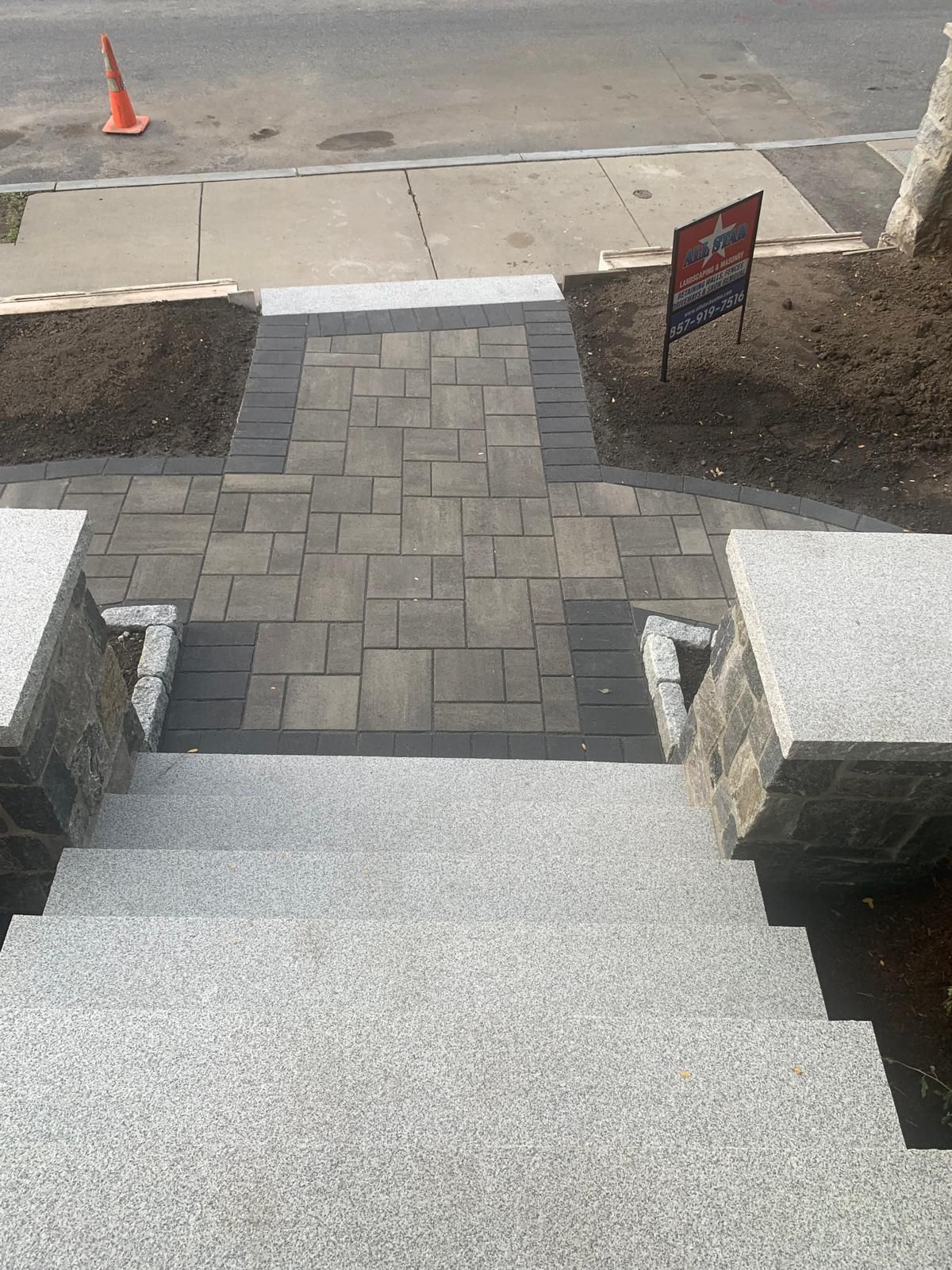 Front steps leading to a paved sidewalk, with a road, traffic cone, and yard sign ahead.