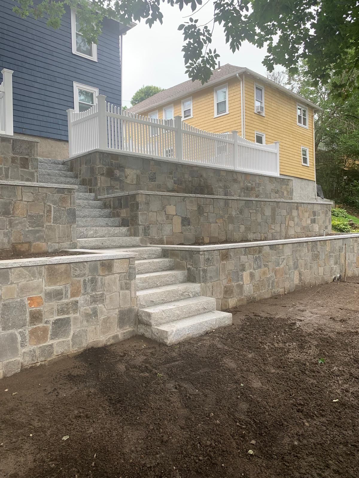 Tiered stone retaining wall with white stairs leading to a raised patio beside a yellow house