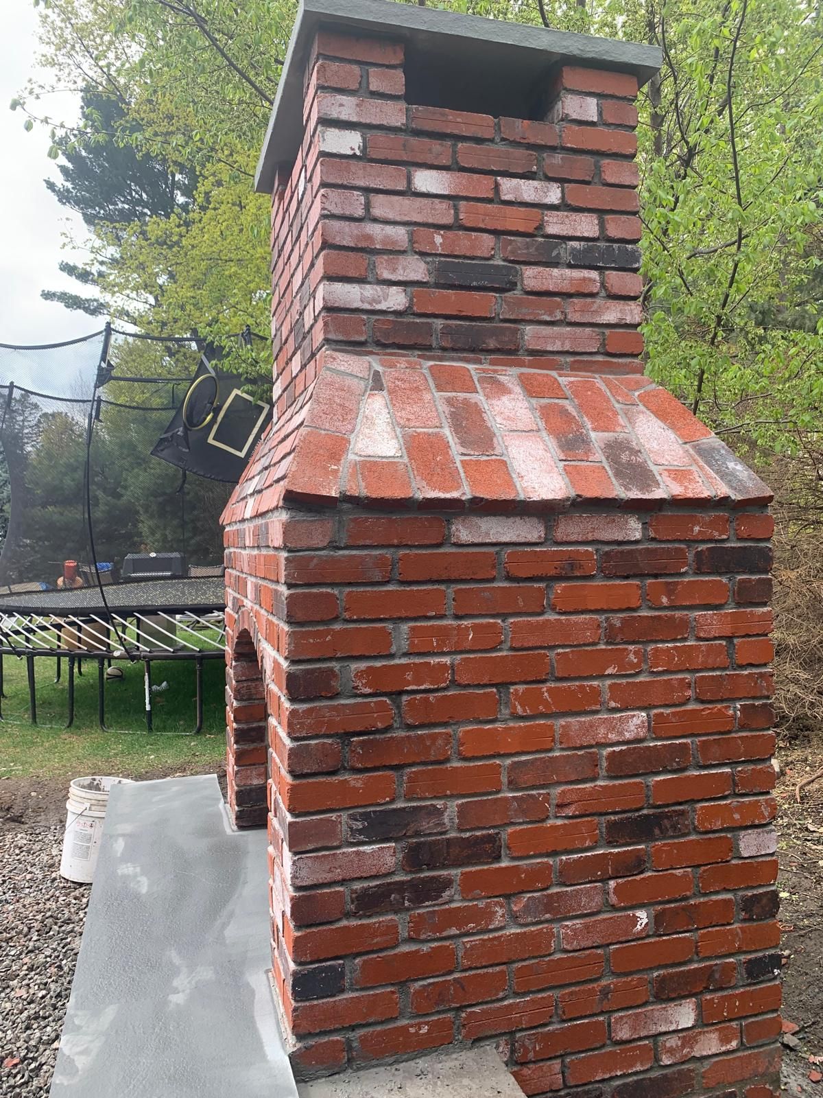 Brick outdoor chimney with a metal cap and sloped flashing, standing on a concrete base in a yard.