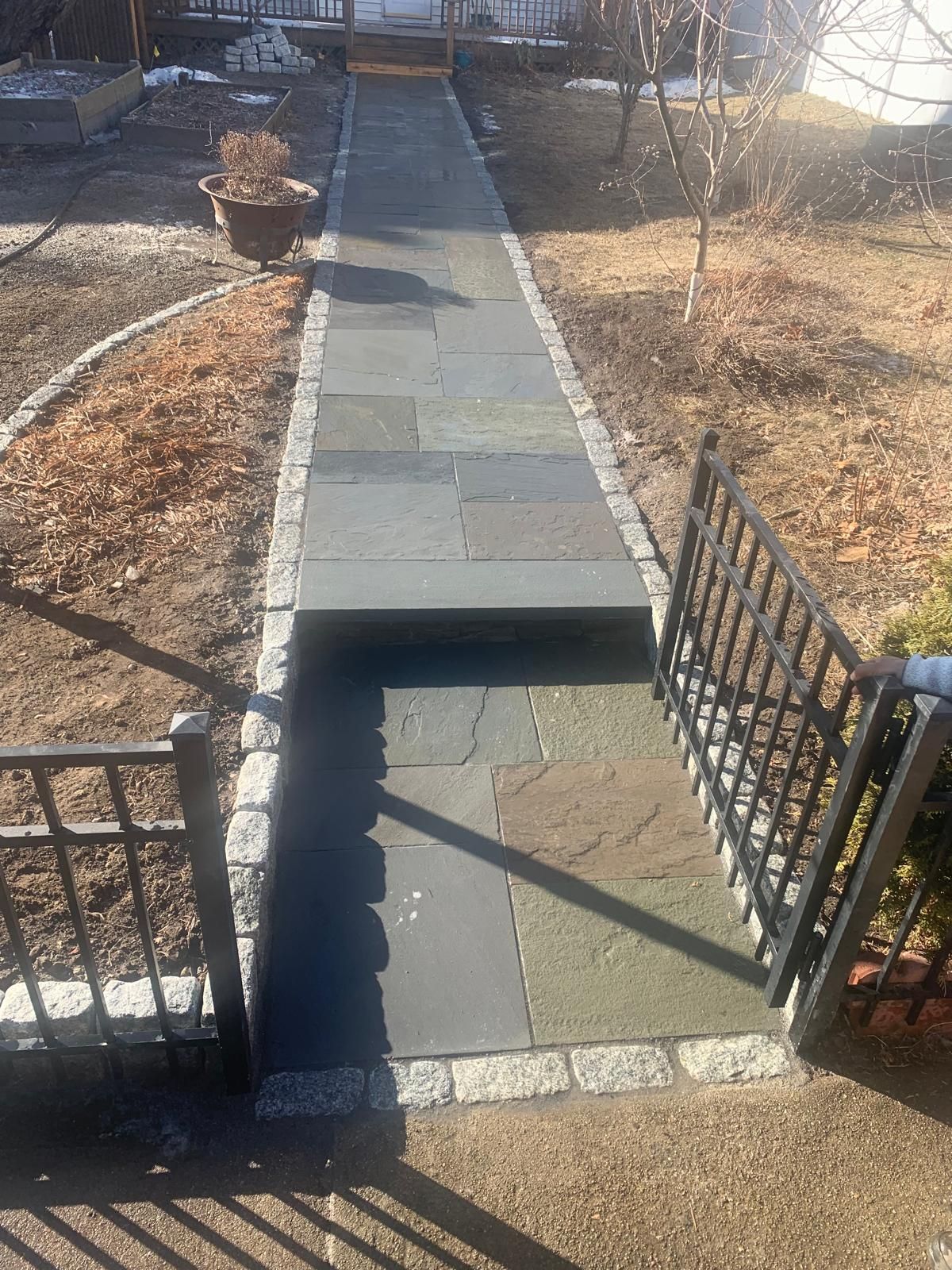 Narrow stone walkway with a raised step gap, bordered by black metal fences and dry landscaping.