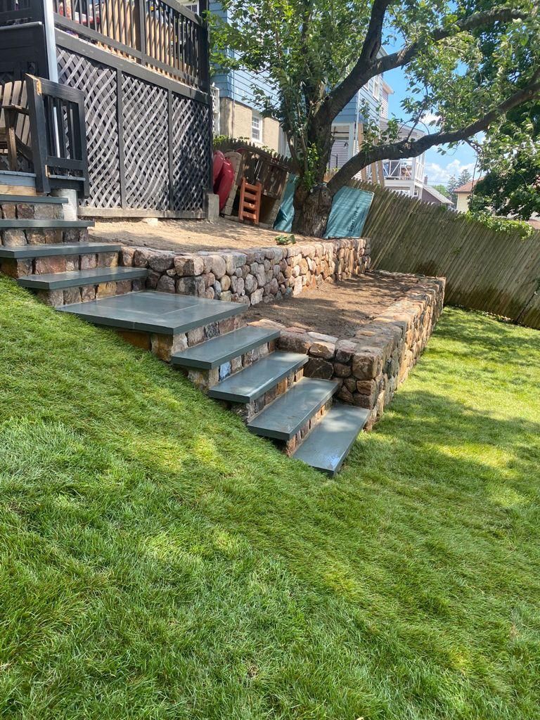 Stone steps and retaining wall on a grassy backyard slope beside a house, with a tree and fence.