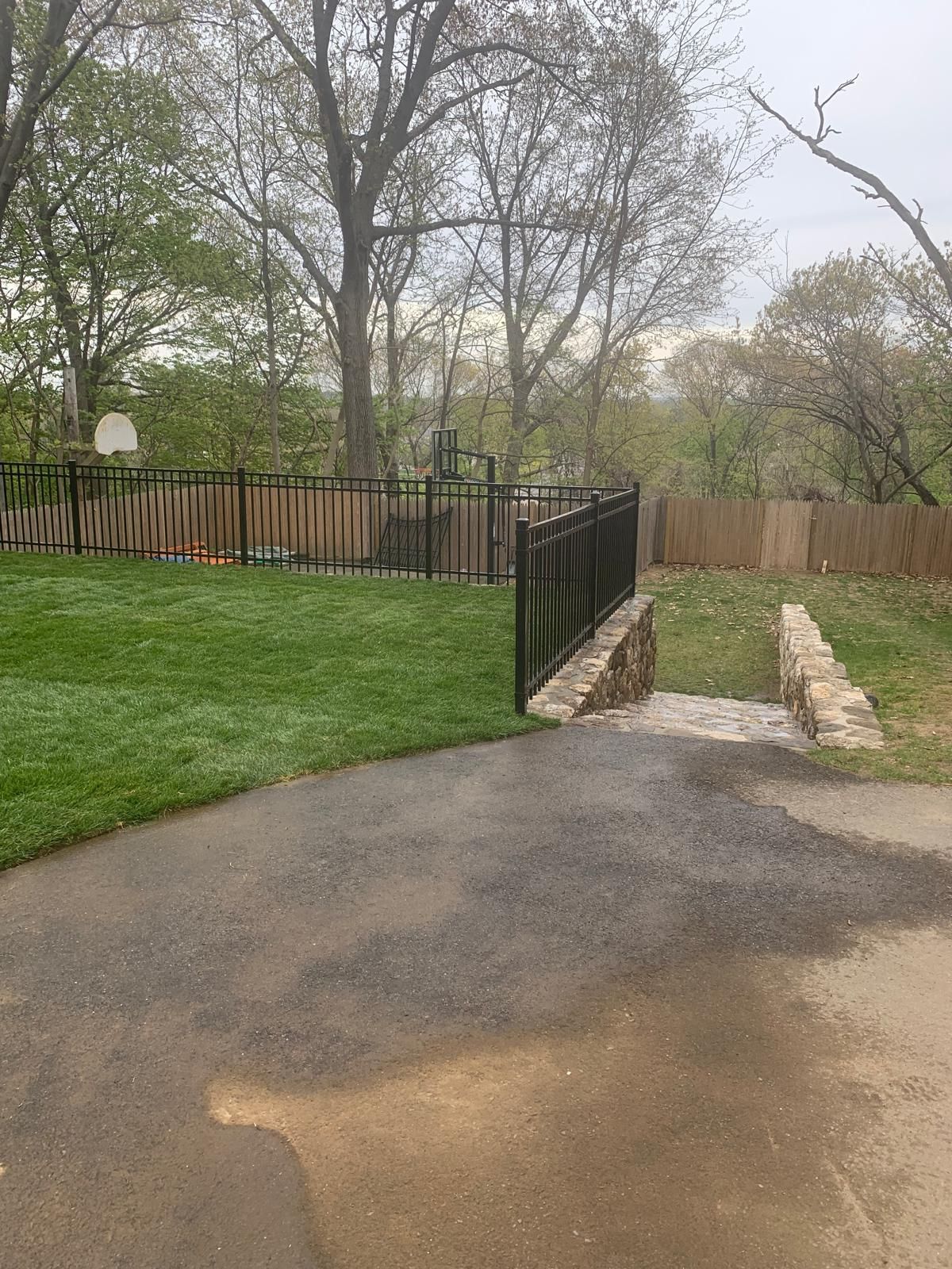 Backyard driveway leading to a fenced gate and stone path, with green lawn and leafless trees.