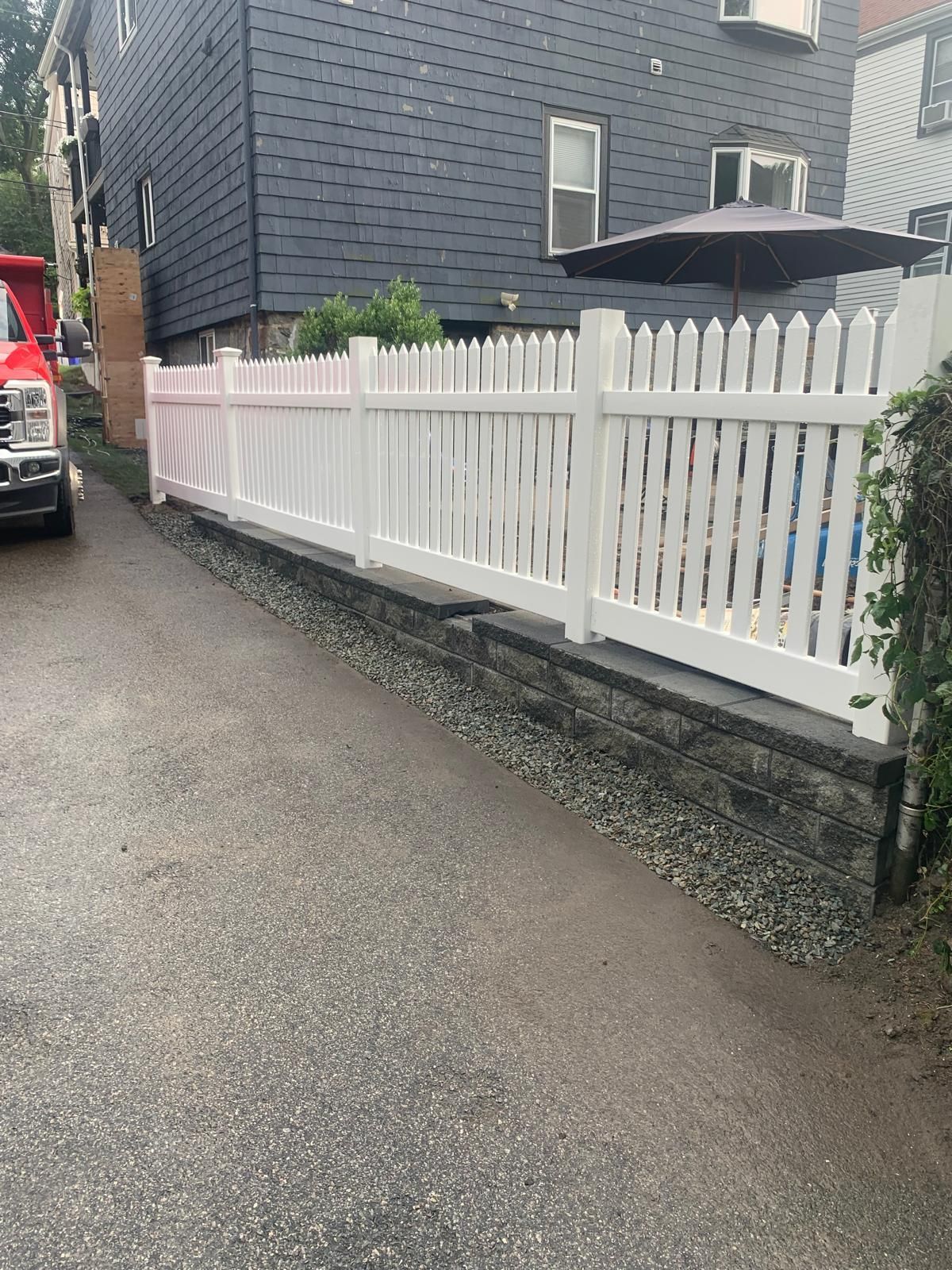 White picket fence along a wet alley beside a gray house, with a parked car and patio umbrella.