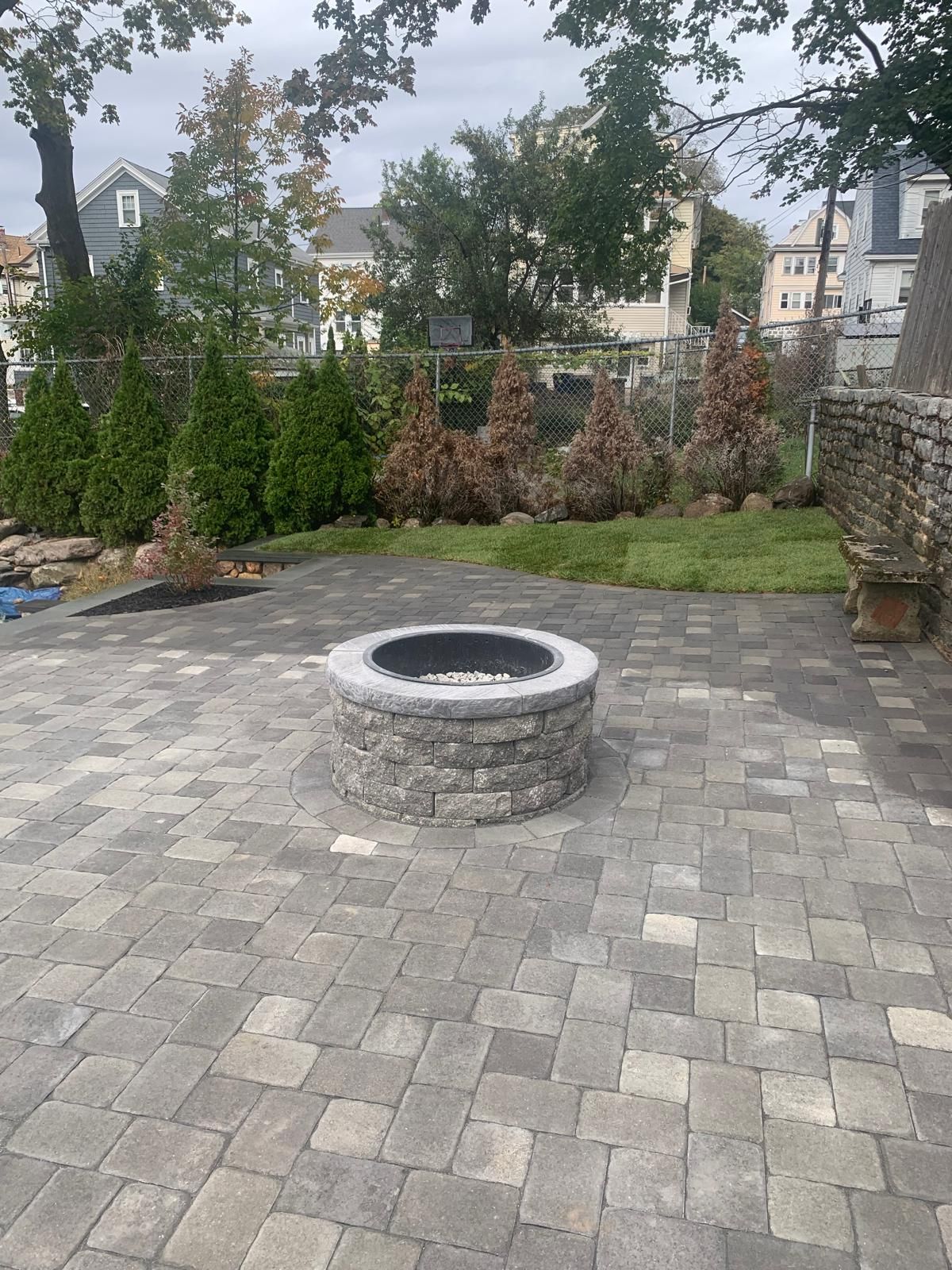 Stone fire pit in a paved backyard patio with a hedge and houses in the background