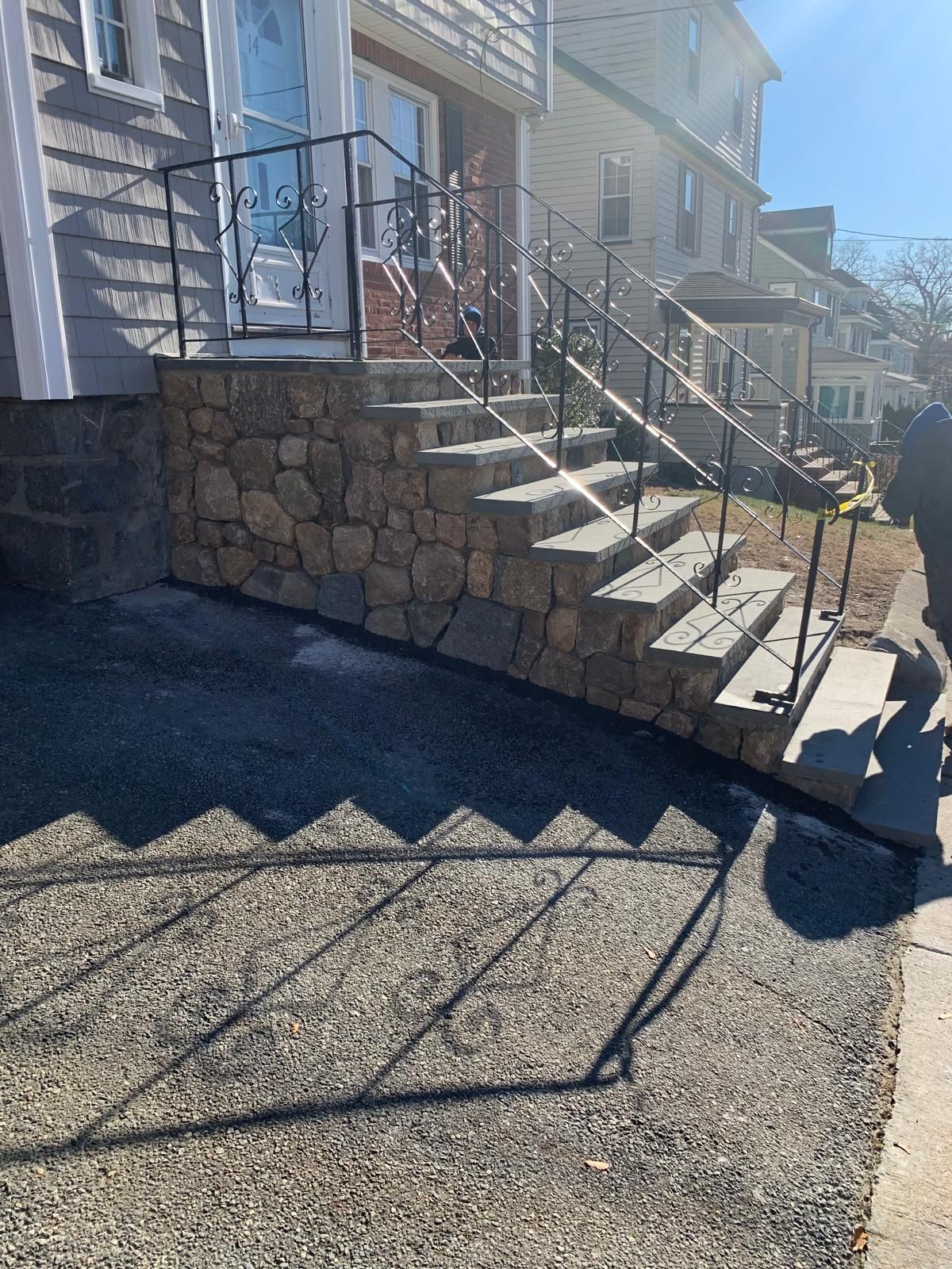 Stone retaining wall with concrete steps and metal railing beside a house, leading down to a gravel driveway.