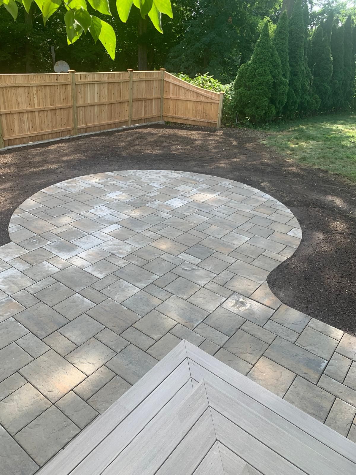 Backyard patio with light pavers, a wooden fence, and surrounding dark soil and greenery.