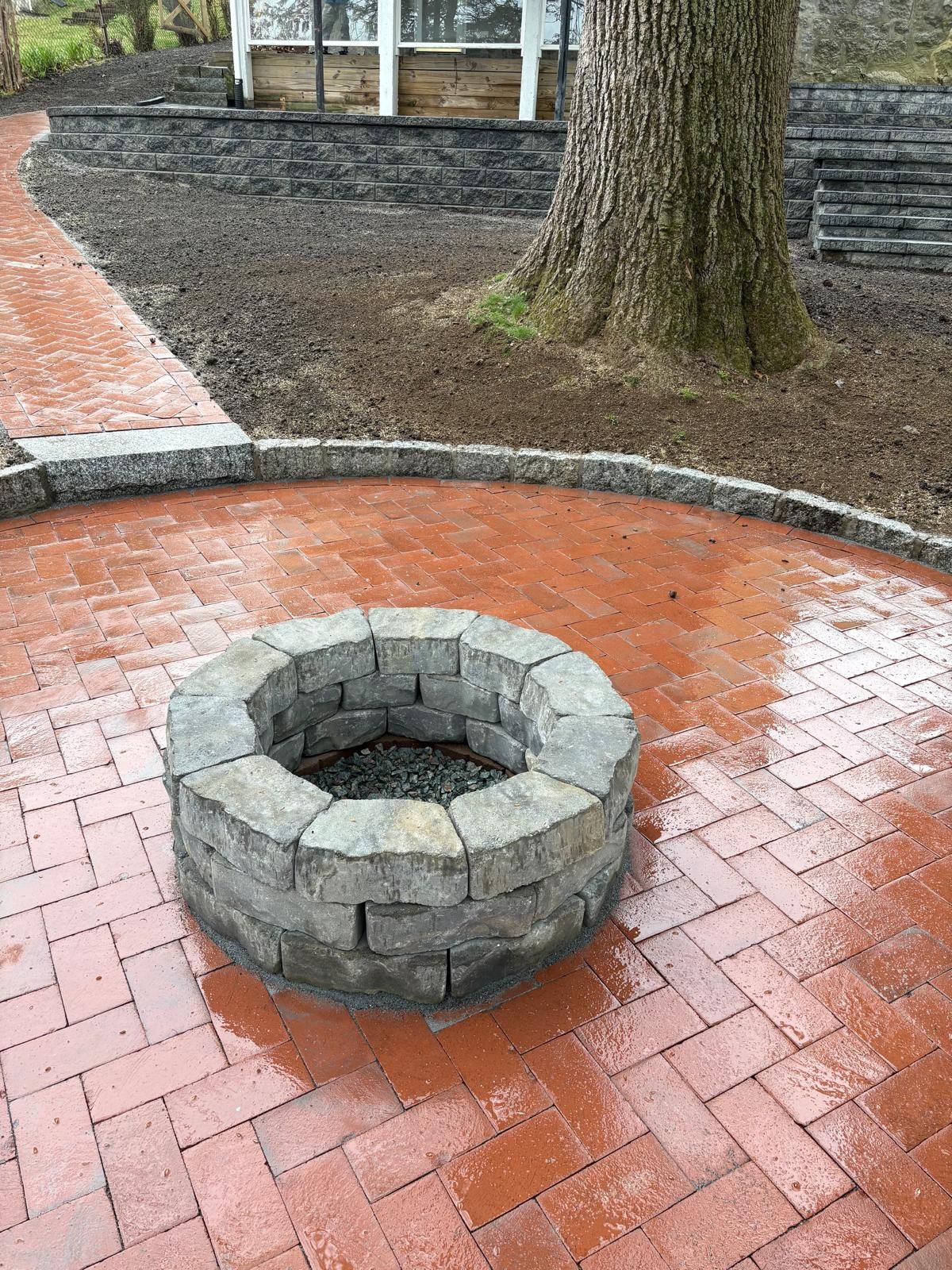 Stone fire pit on a red brick patio beside a tree in a park.