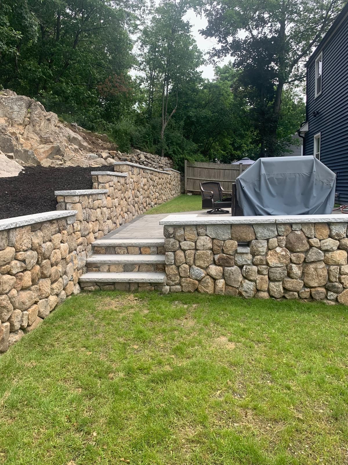 Stone patio with steps, retaining walls, grass lawn, and a covered grill beside a house