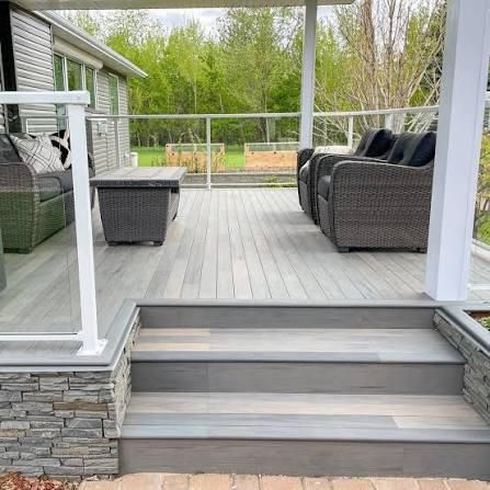 Covered backyard deck with gray steps, wicker seating, and glass railing overlooking a green yard