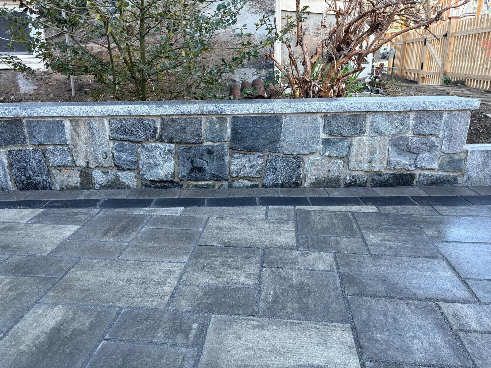 Stone retaining wall beside a gray paved patio with shrubs and trees behind it