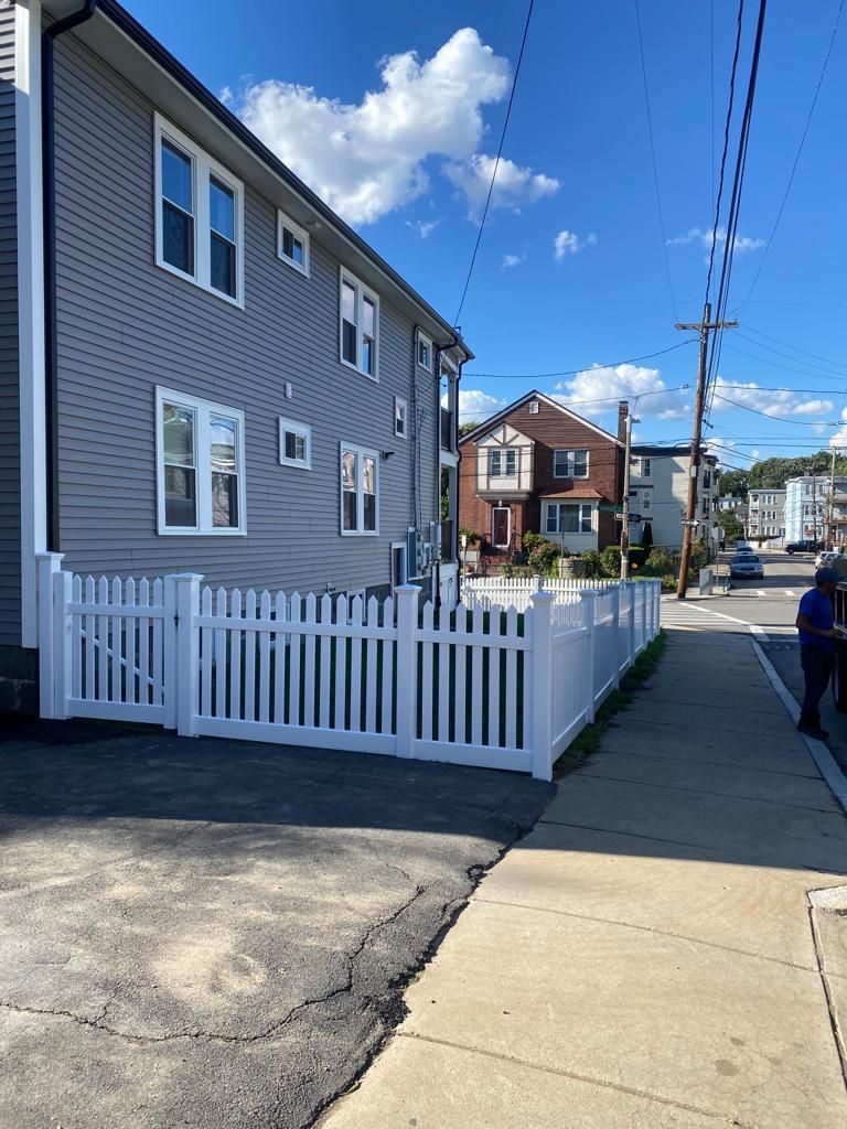 Residential street with gray house, white fence, and sidewalk under a bright blue sky