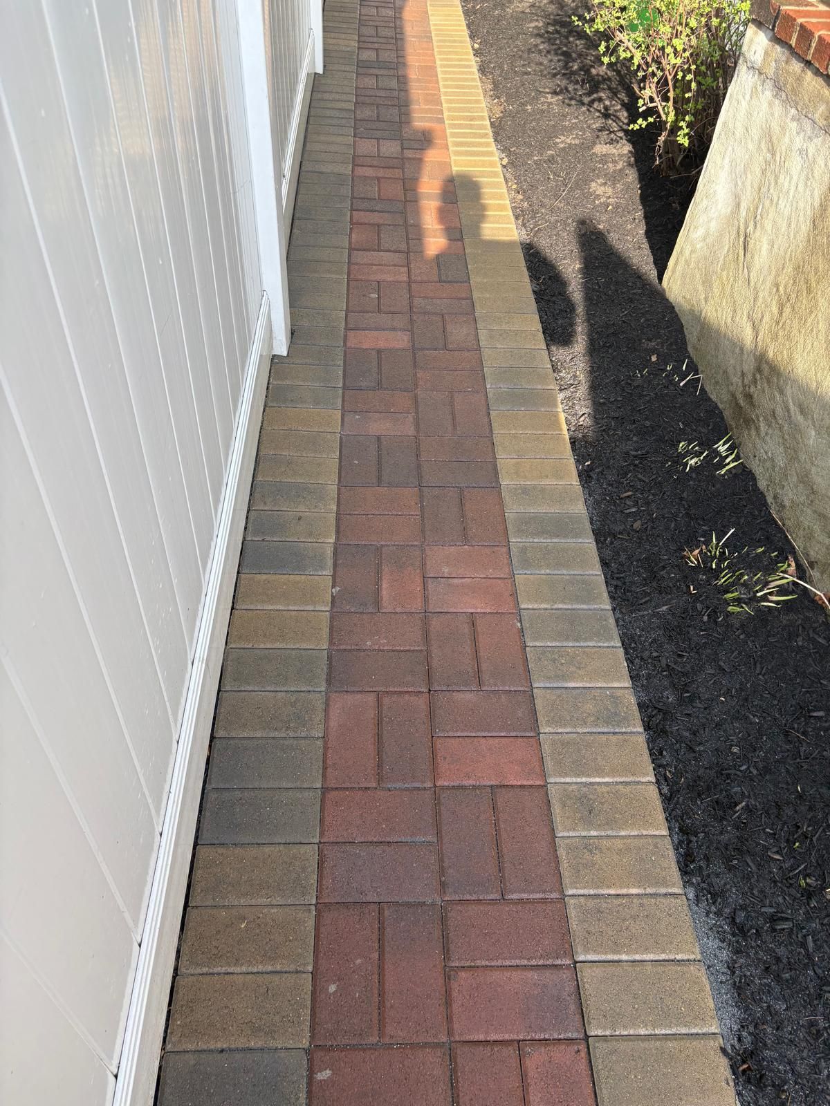 Narrow brick pathway between a white fence and a wall, with a shadow cast along the right side.
