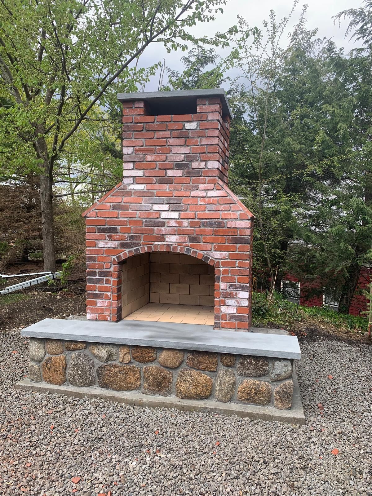 Outdoor brick fireplace with a stone base and chimney in a wooded backyard.