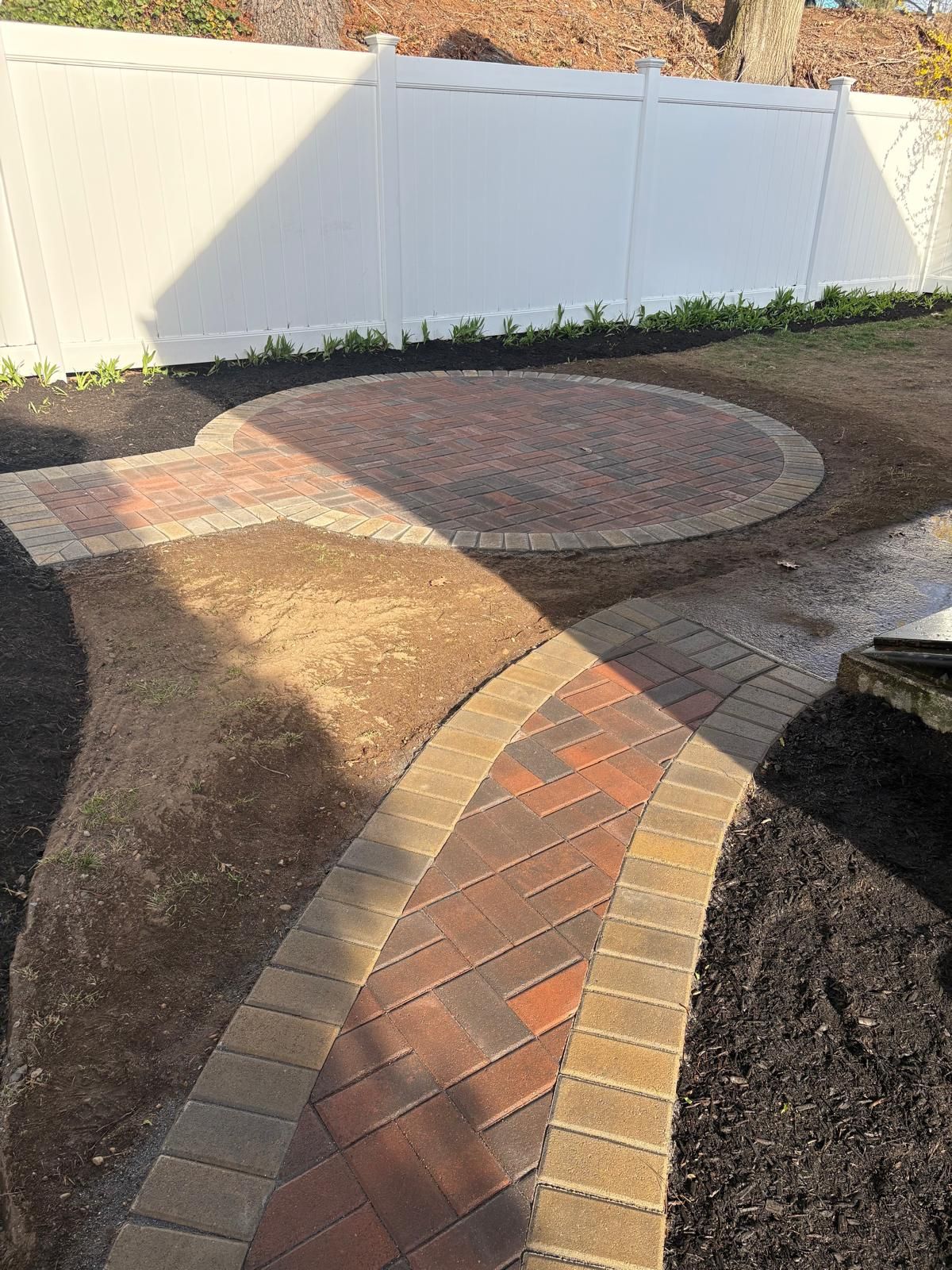 Curved brick walkway leading to a circular patio in a fenced backyard with bare garden beds.