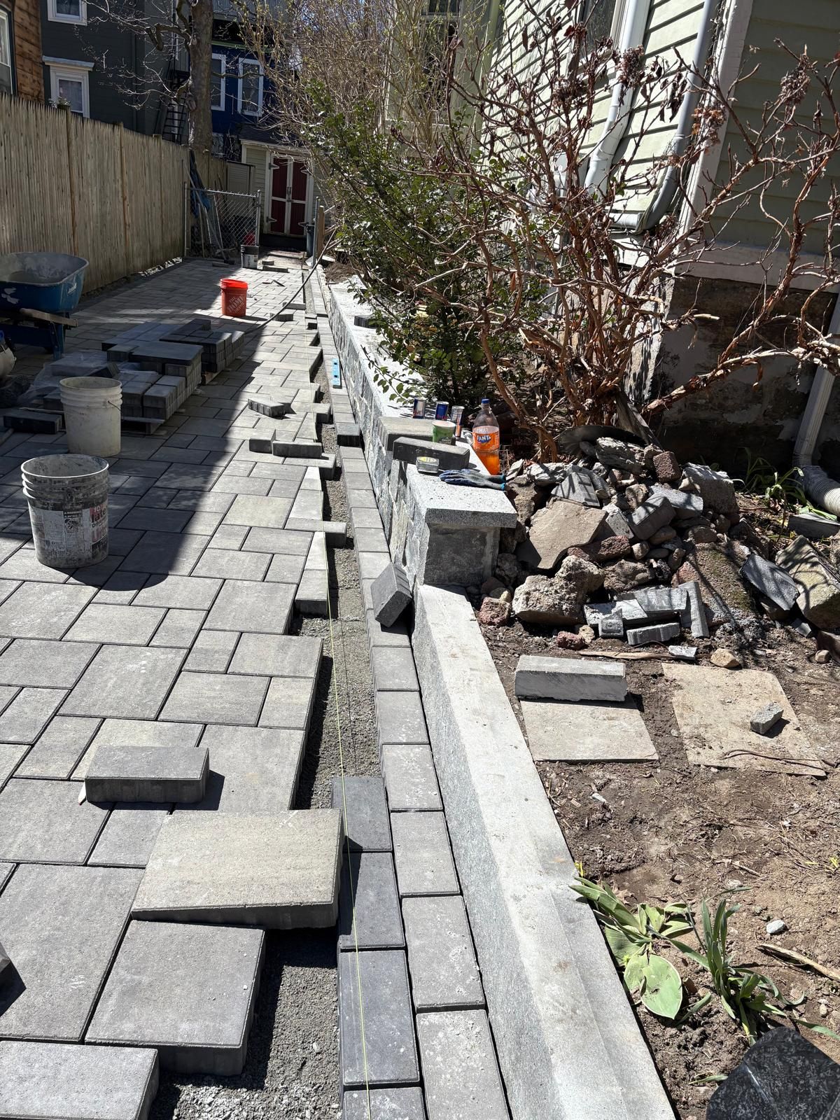 Partially paved backyard walkway with gray pavers, buckets, and stacked stones beside a building and fence