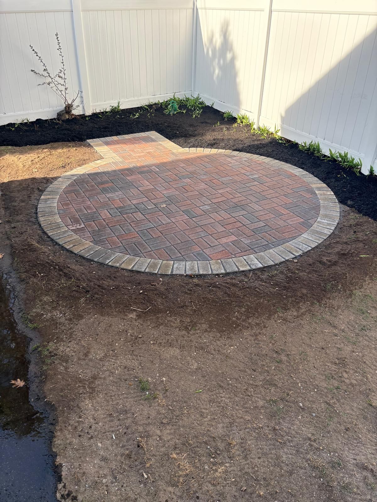 Circular brick patio in a fenced backyard with bare soil around it and a small paved step area.