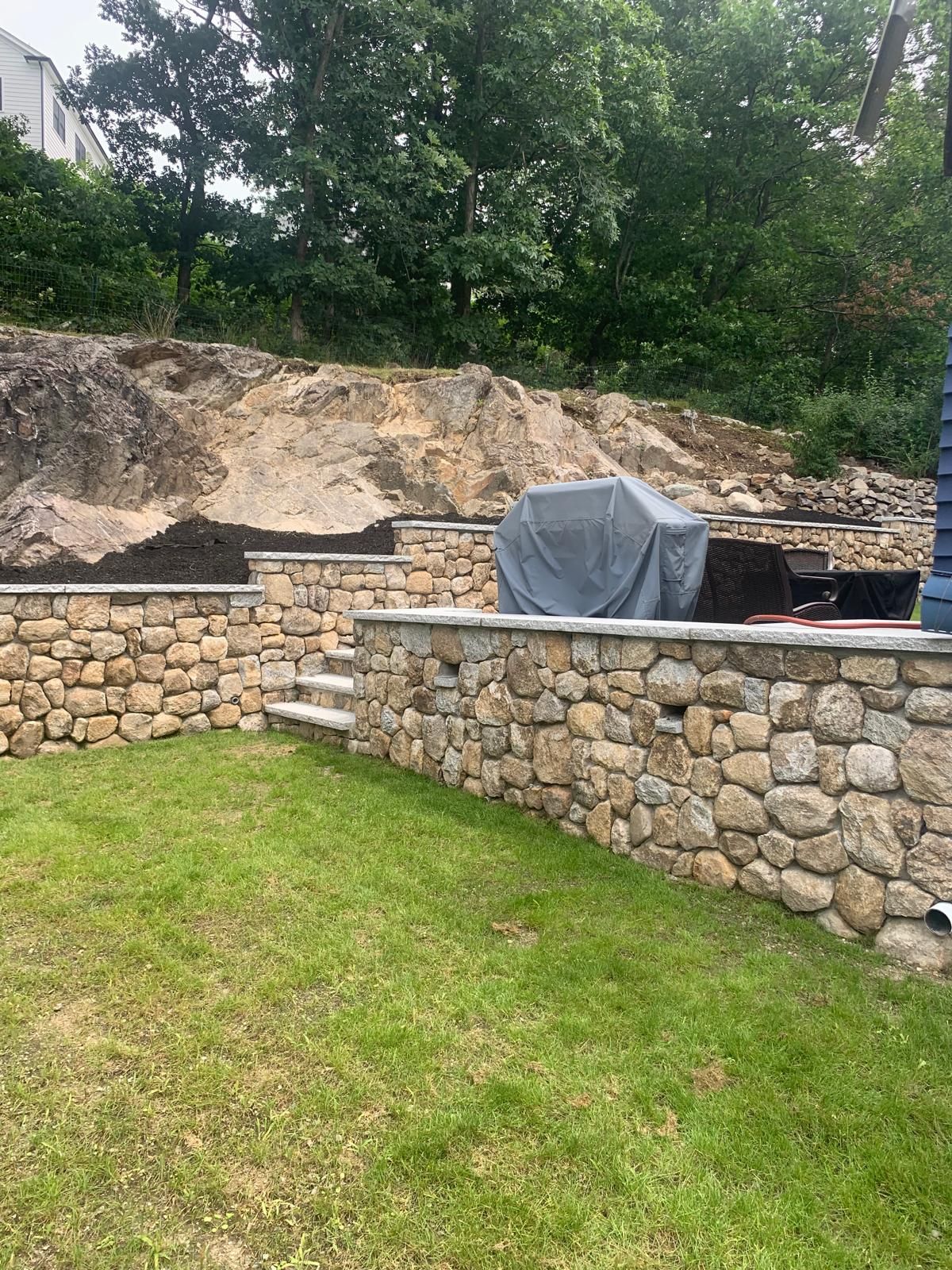 Backyard stone retaining walls with a covered grill and grassy lawn below a wooded slope