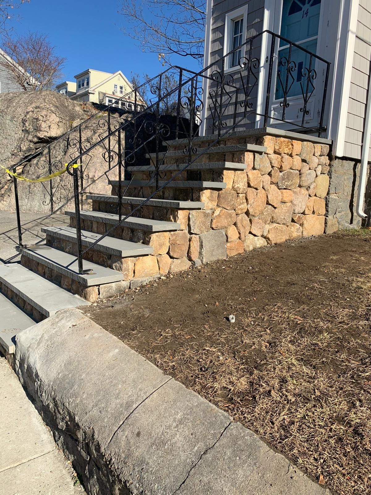 Front steps with black railing beside a stone retaining wall and sloped yard beside a house.