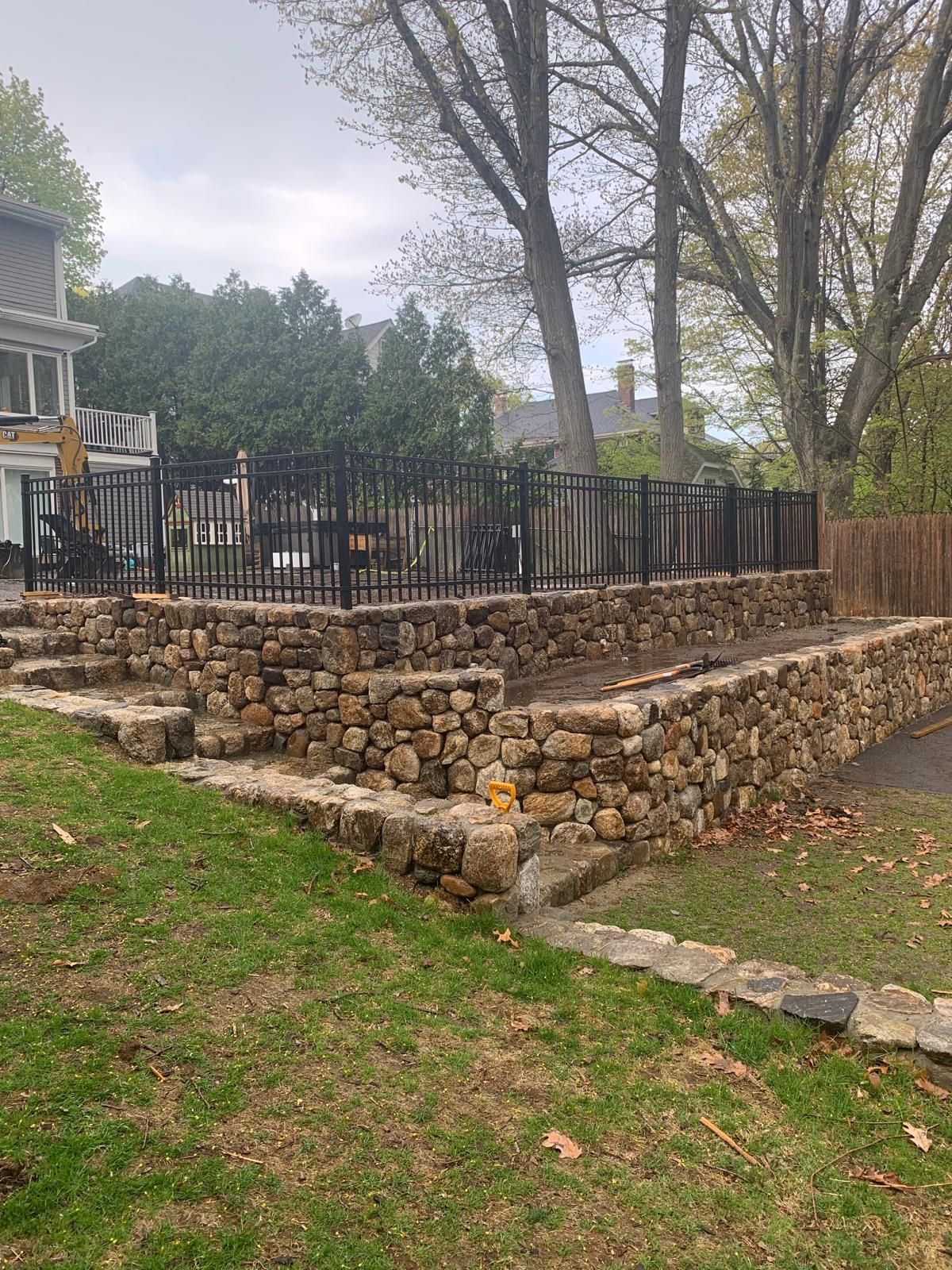 Stone retaining wall and black fence bordering a raised patio in a grassy backyard, with trees overhead.