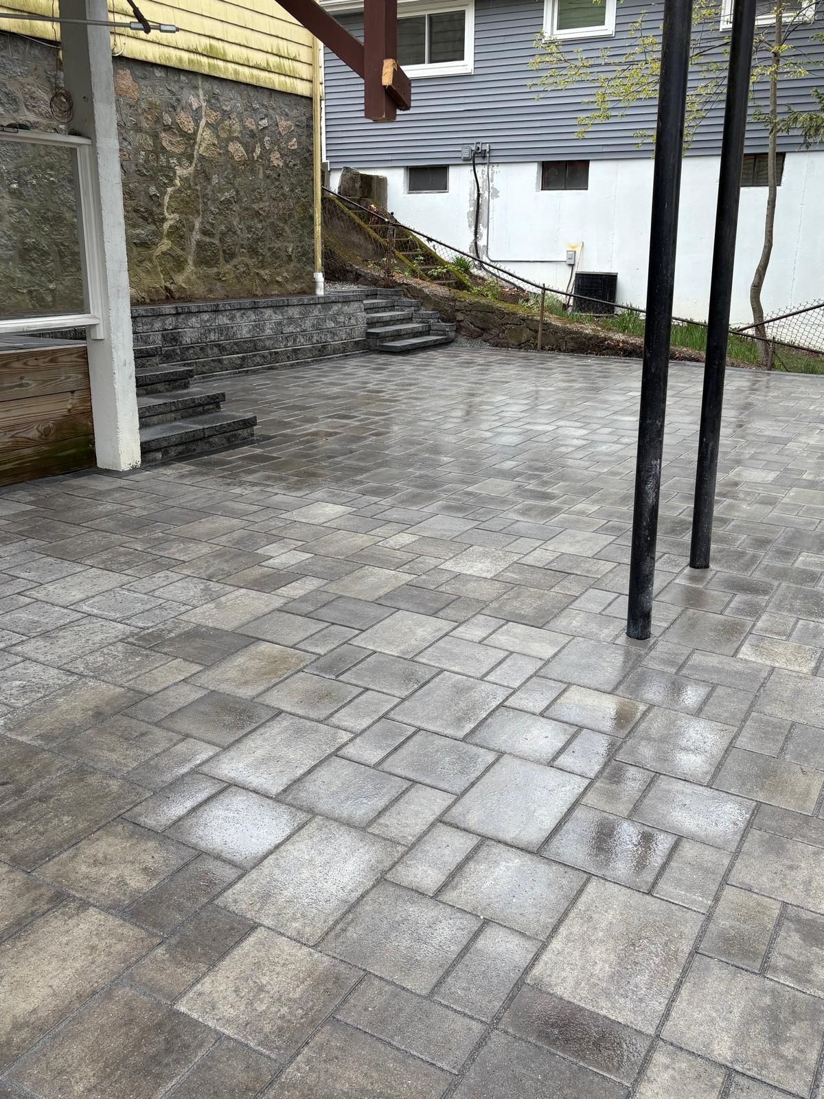 Outdoor stone patio beside a house, with steps, retaining wall, and two black support poles.