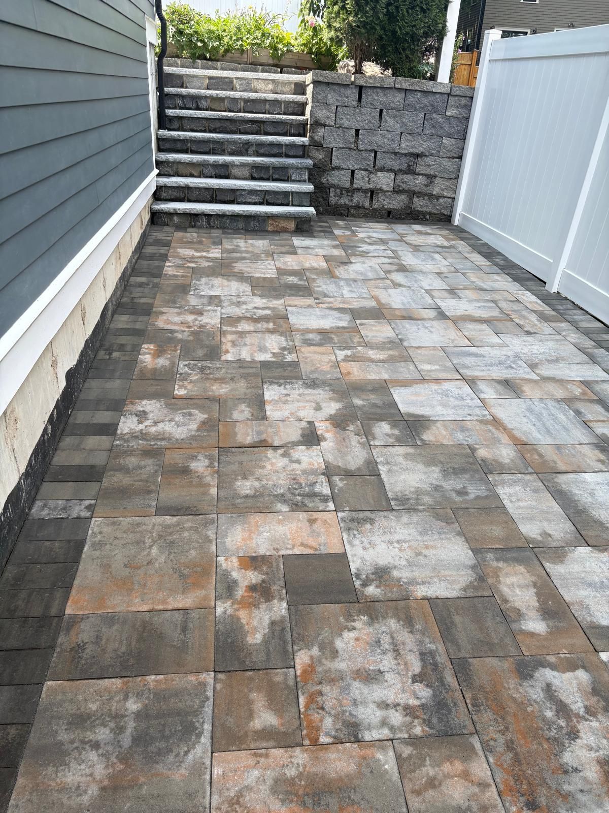 Stone-paved backyard patio with steps leading to a raised area, bordered by a fence and house siding.
