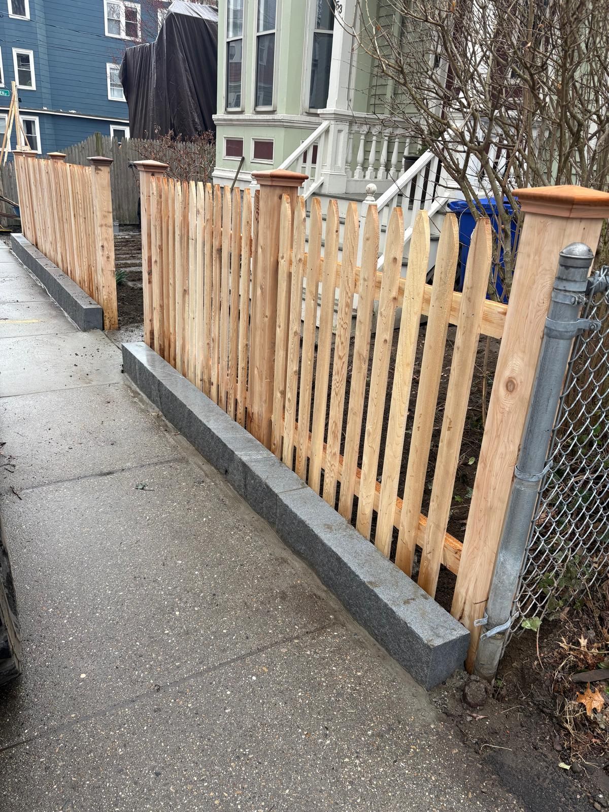 Wooden fence along a wet sidewalk beside a chain-link gate and residential houses