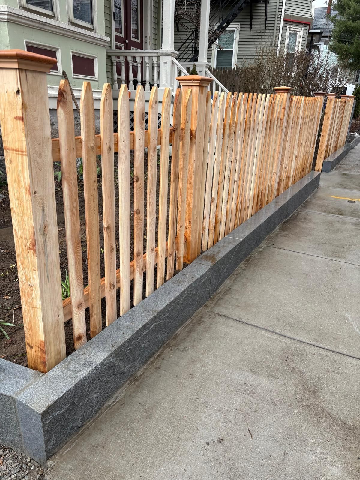 Wooden picket fence with concrete base along a sidewalk beside a residential building.