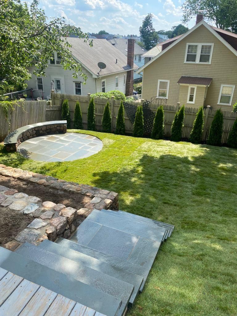Backyard patio with stone steps, curved retaining wall, lawn, and evergreen privacy shrubs, houses beyond