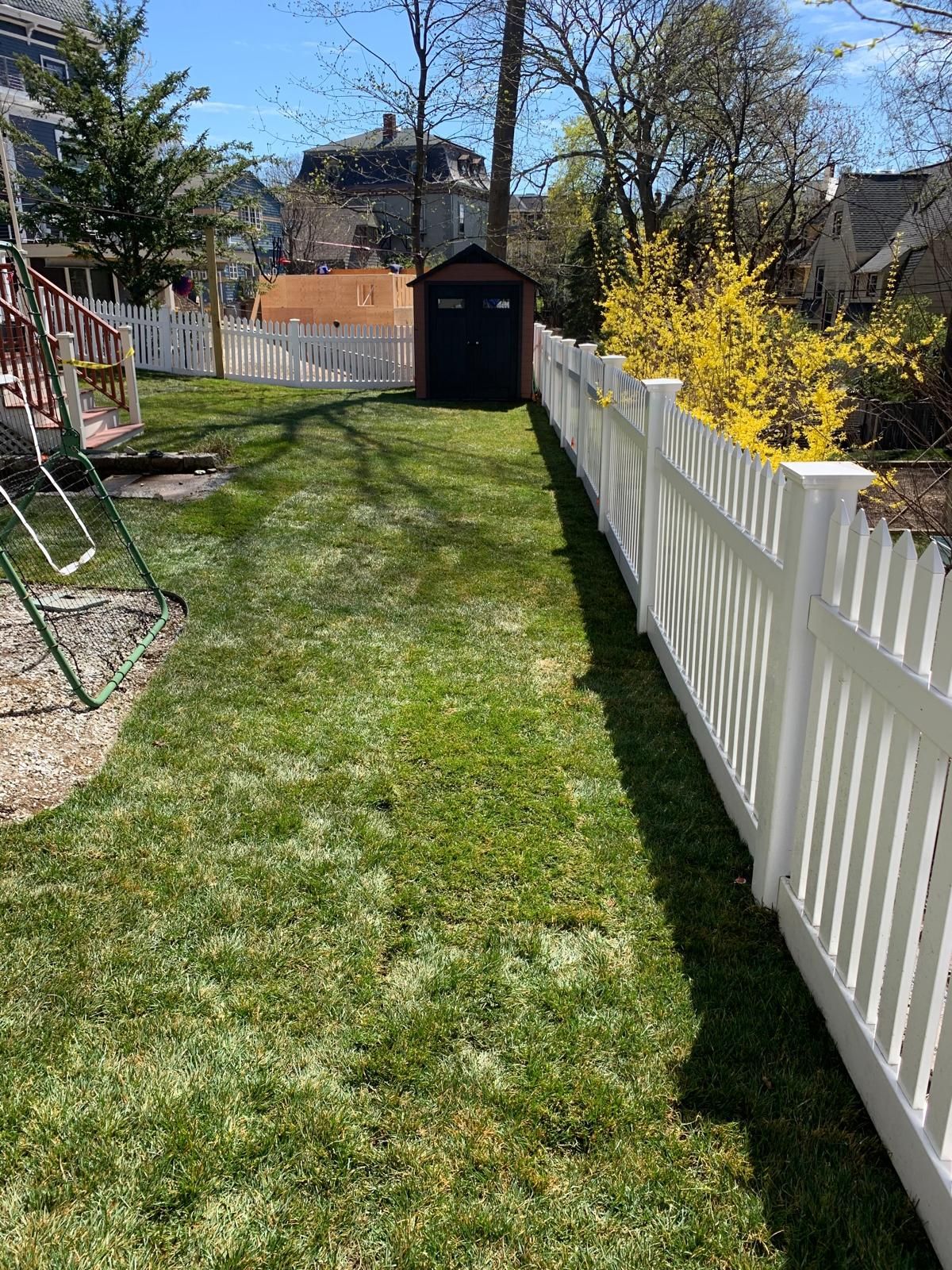 Narrow backyard lawn between a white fence and house, with a shed at the far end on a sunny day