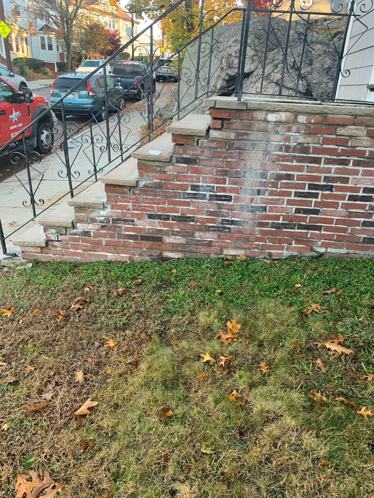 Brick front stoop with wrought-iron railing beside a grassy yard and sidewalk with parked cars on a street