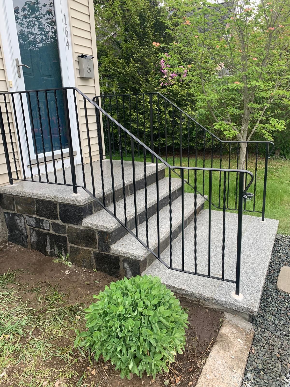 Front steps with black railing leading to a blue door beside a stone house and green shrubs