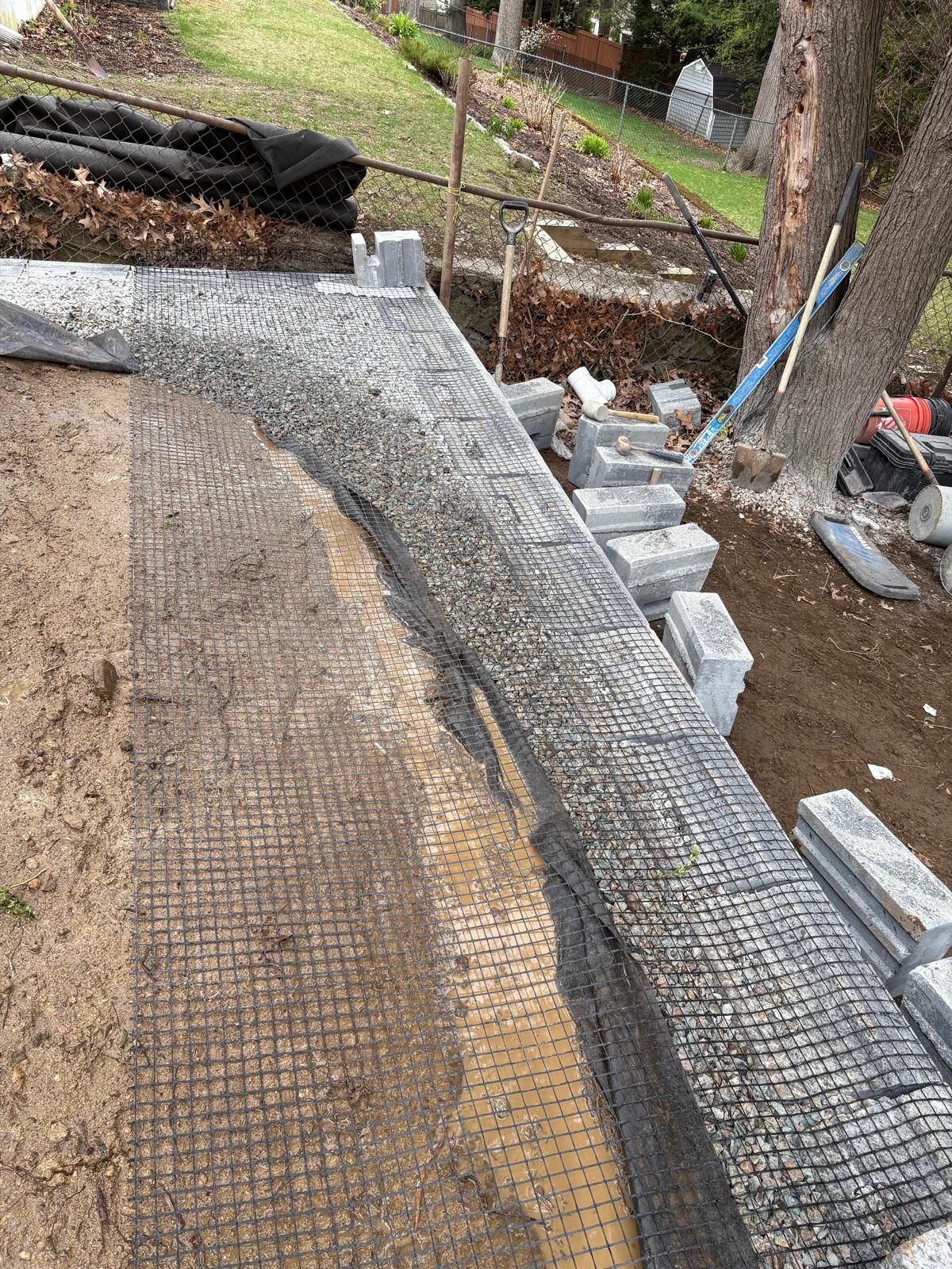 Curved concrete walkway under construction with wire mesh, gravel, and stacked pavers beside a wooded yard
