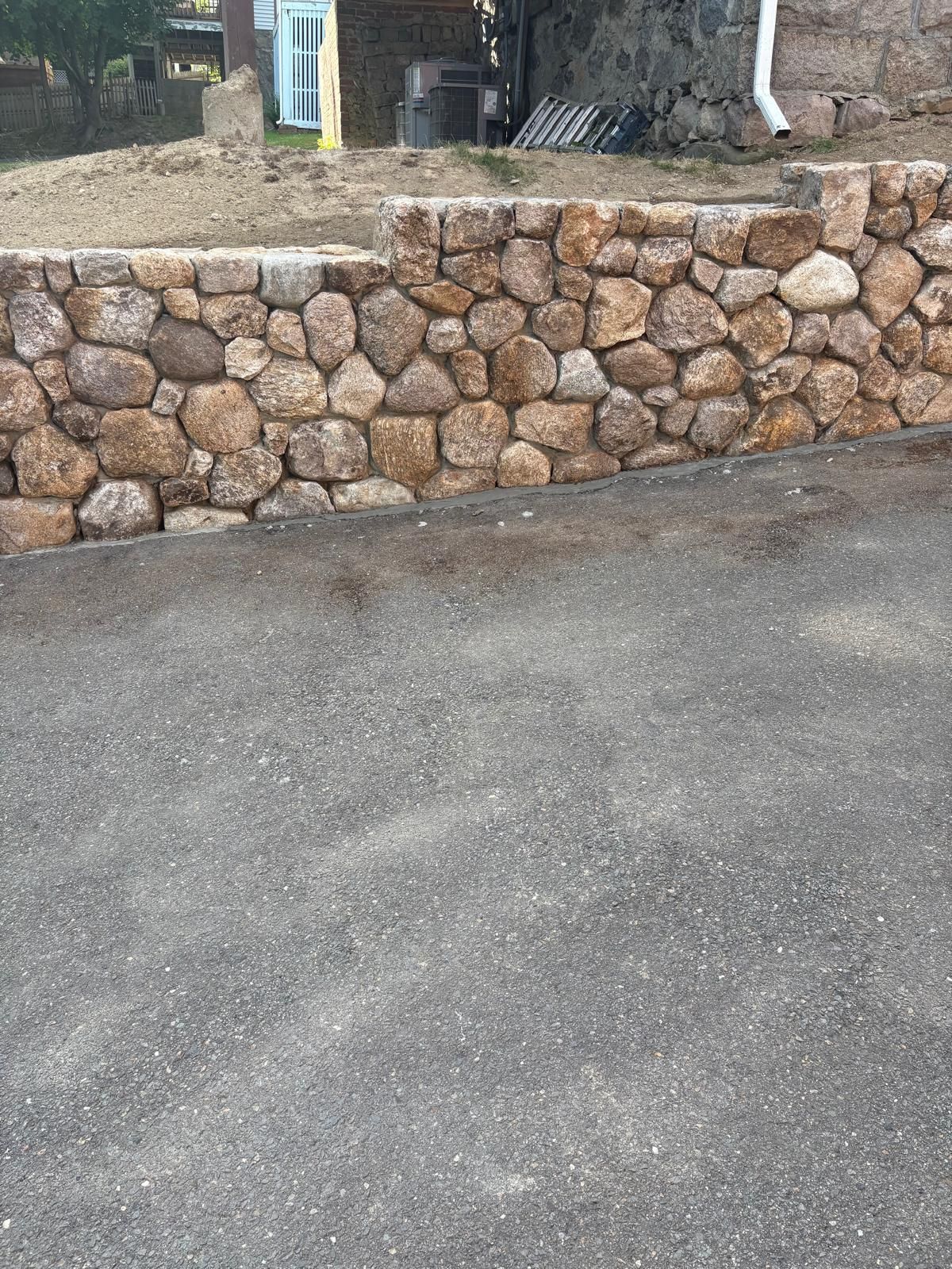Stone retaining wall beside a paved road, with a building and trees in the background.