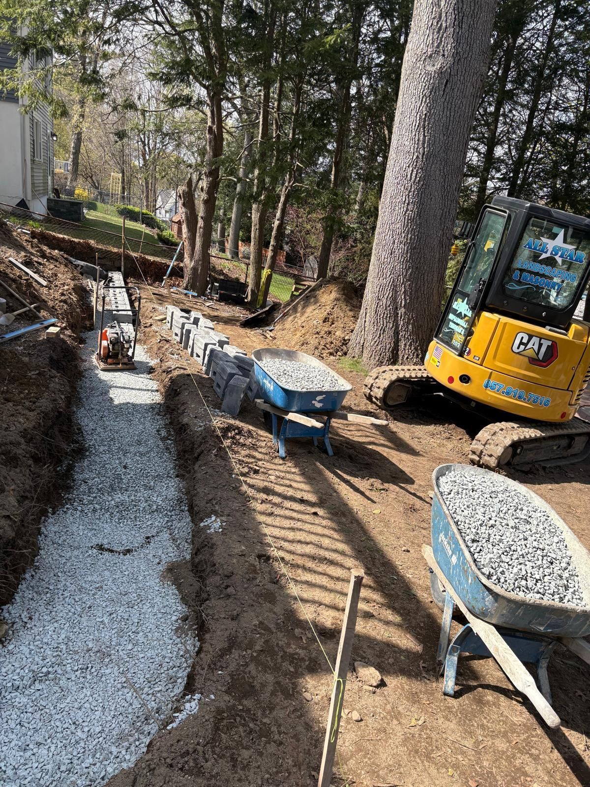 Construction site with gravel path, wheelbarrows, and a yellow excavator beside a wooded area