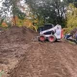 White skid-steer loader on a muddy dirt path beside a large soil pile in a wooded area