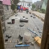 Brick patio under construction with paving stones, tools, and workers in the background