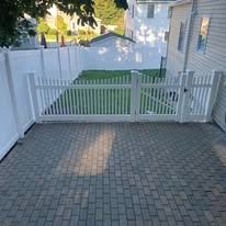 White vinyl gate and fence opening onto a brick-paved patio beside a house