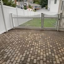 Brick-paved patio facing a white picket gate and fence, with a grassy yard beyond