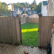 Backyard view through a wooden gate, showing green grass and neighboring houses beyond a fence.
