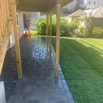 Covered backyard patio with stone pavers beside a green lawn and garden, lit by sunlight