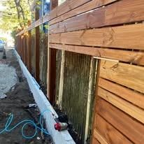 Long wooden fence beside a dirt path, with a white concrete base and blue hose along the ground.