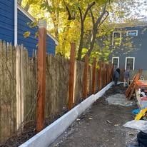 Backyard fence under construction beside a blue house, with white base trim and tools along the narrow path