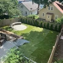 Backyard with green lawn, patio deck, round fire pit, and houses beyond a privacy fence