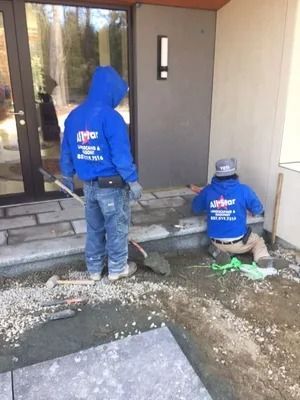 Two workers in blue hoodies kneel and stand by a front porch, laying a walkway beside a house door.